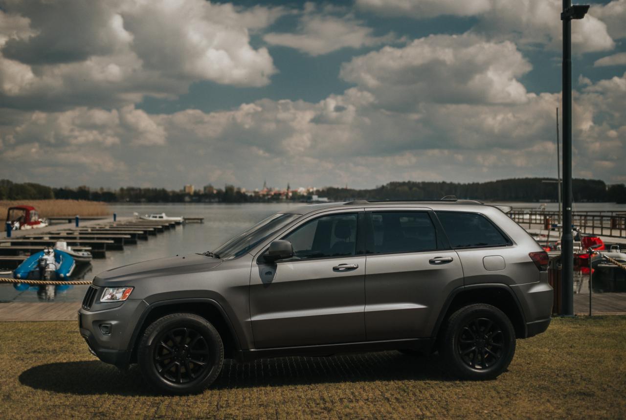 Szary Jeep Grand Cherokee z czarnymi felgami, zaparkowany na trawie obok jeziora z pomostami i łodziami w tle, pochmurne niebo.