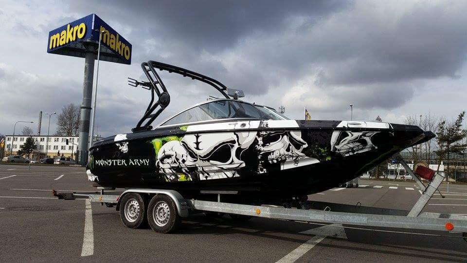 Czarno-biała łódź z grafiką czaszek i napisem 'Monster Army' na przyczepie, na tle parkingu i budynku Makro.