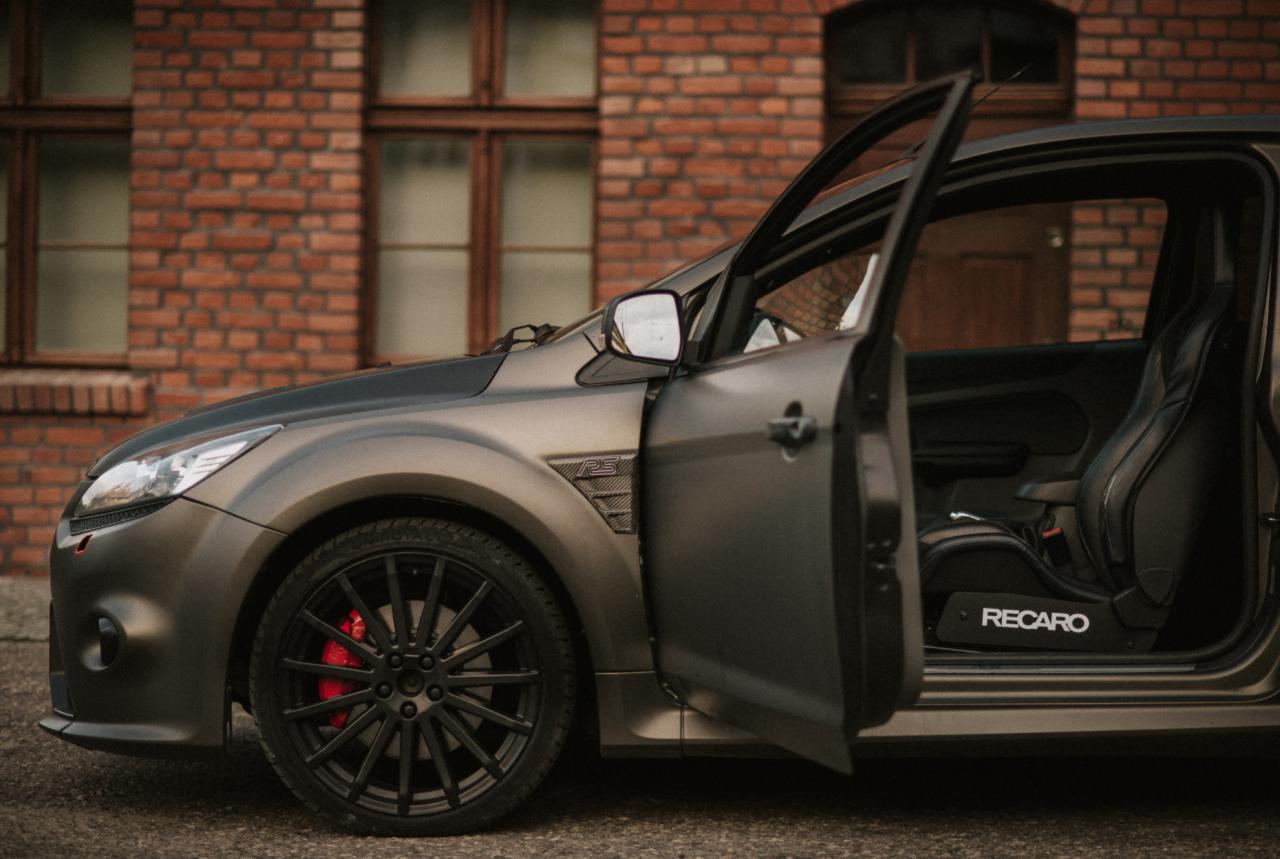 Matowy, szary Ford Focus RS z otwartymi drzwiami, widoczne czerwone zaciski hamulcowe i fotele Recaro, na tle ceglanej ściany budynku.
