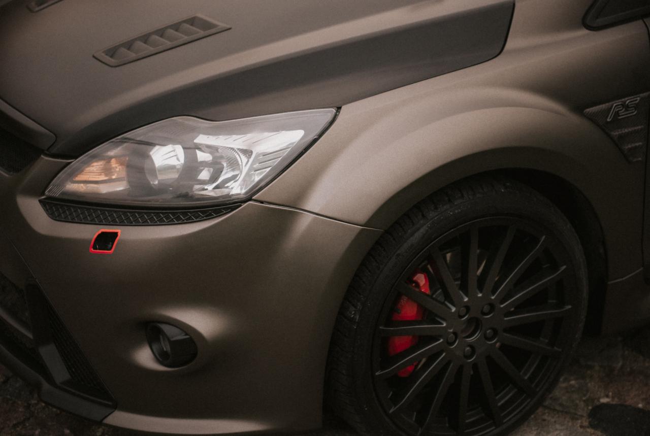 Matowy, oliwkowy Ford Focus RS z czarnymi felgami i czerwonymi zaciskami hamulcowymi, widok na przedni reflektor i fragment maski z wlotami powietrza.