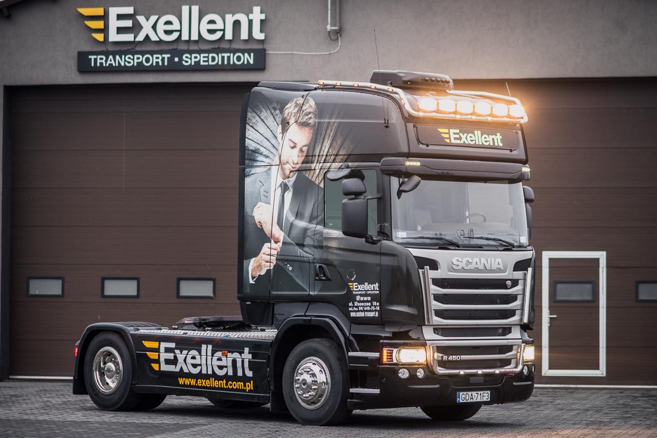 Czarny ciągnik siodłowy Scania z grafiką mężczyzny w garniturze na kabinie, logo firmy Exellent Transport Spedition na boku i dachu, zaparkowany przed budynkiem firmy z brązową bramą garażową.