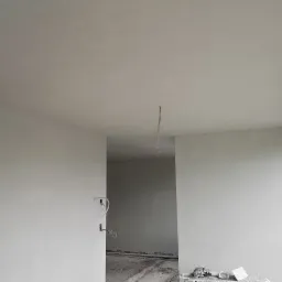 Świeżo otynkowane ściany i sufit w jasnym pomieszczeniu z widocznymi przewodami elektrycznymi i fragmentem okna, gotowe do dalszych prac wykończeniowych.