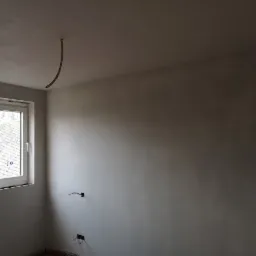 Wnętrze pokoju po nałożeniu gładzi, widoczne wyprowadzone kable elektryczne i okno z widokiem na dach.