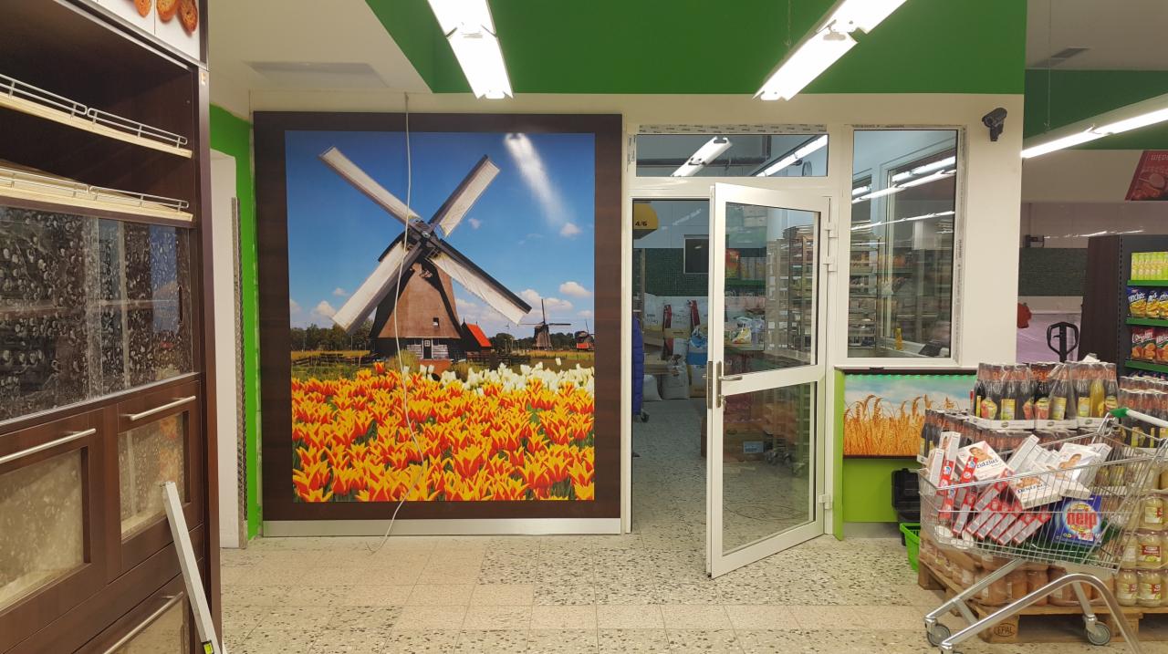 Wnętrze sklepu spożywczego z dekoracyjną fototapetą przedstawiającą holenderski krajobraz z wiatrakiem i polem tulipanów, umieszczoną na ścianie w pobliżu wejścia.