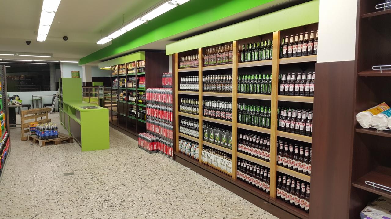 Wnętrze sklepu spożywczego z regałami pełnymi butelek piwa, jasnymi, zielonymi akcentami kolorystycznymi i kasą w tle.