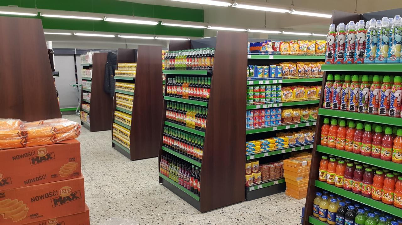 Wnętrze sklepu spożywczego z regałami wypełnionymi produktami, widok na alejki między regałami, na pierwszym planie kartony z przekąskami.