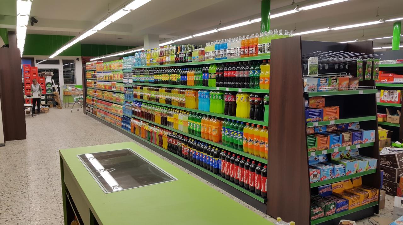Wnętrze sklepu spożywczego z pełnymi półkami napojów i słodyczy, widok z perspektywy lady chłodniczej z zielonym wykończeniem.