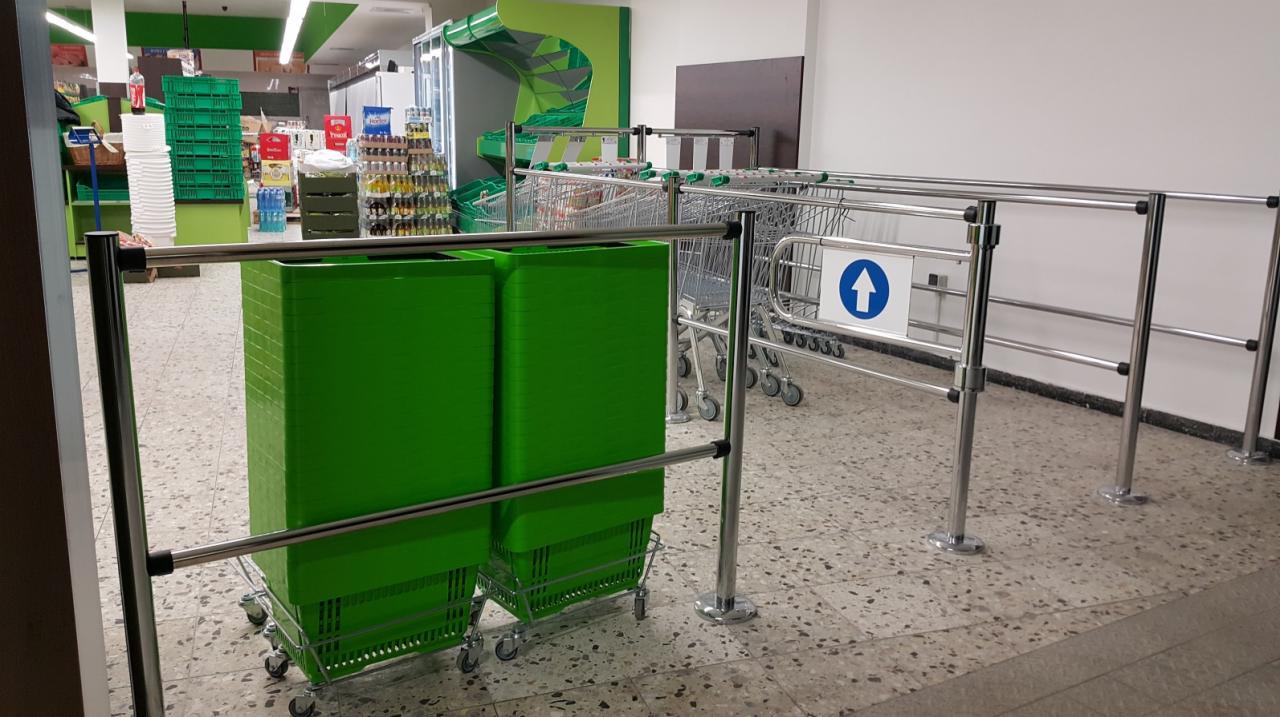 Wnętrze sklepu z widocznymi dwoma wózkami z zielonymi pojemnikami, metalowe barierki z niebieską strzałką wskazującą kierunek oraz wózki sklepowe w tle.