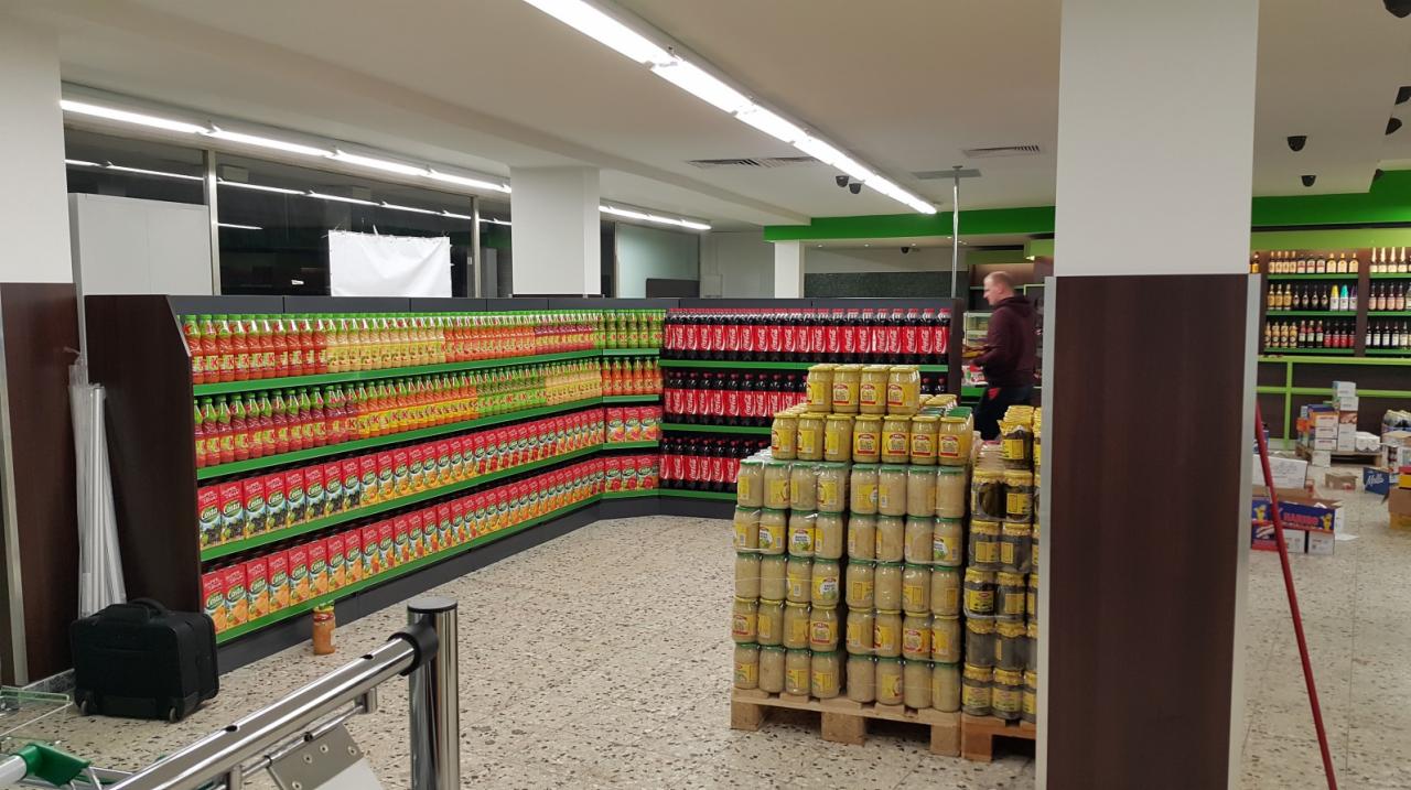 Półki sklepowe z napojami i słoikami konserw, widoczne oświetlenie sufitowe i fragment wózka sklepowego.