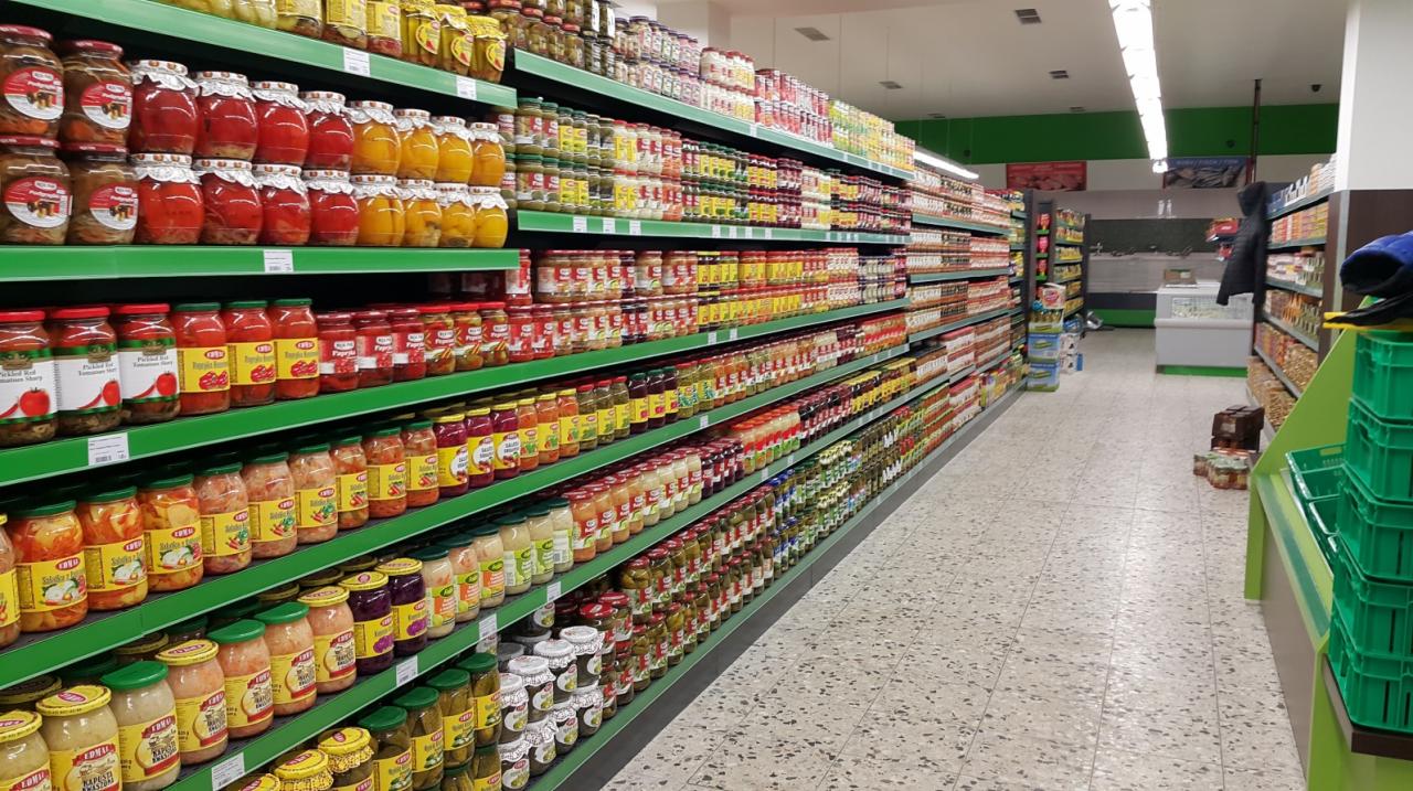 Długi regał sklepowy z zielonymi półkami wypełniony słoikami z marynowanymi warzywami i przetworami, widok z perspektywy alejki sklepowej.