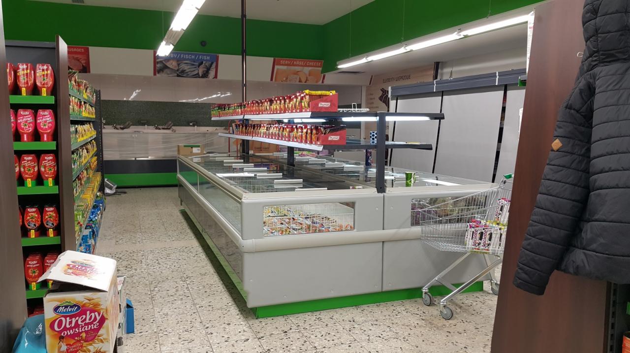 Wnętrze sklepu spożywczego z rzędami przeszklonych lad chłodniczych, regałami z ketchupem i płatkami owsianymi Melvit, widoczne wózki sklepowe i wisząca kurtka zimowa.