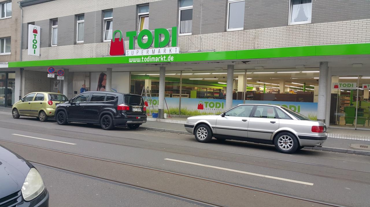 Fasada supermarketu Todi z zielonym logo i reklamą zewnętrzną, zaparkowane samochody na ulicy.