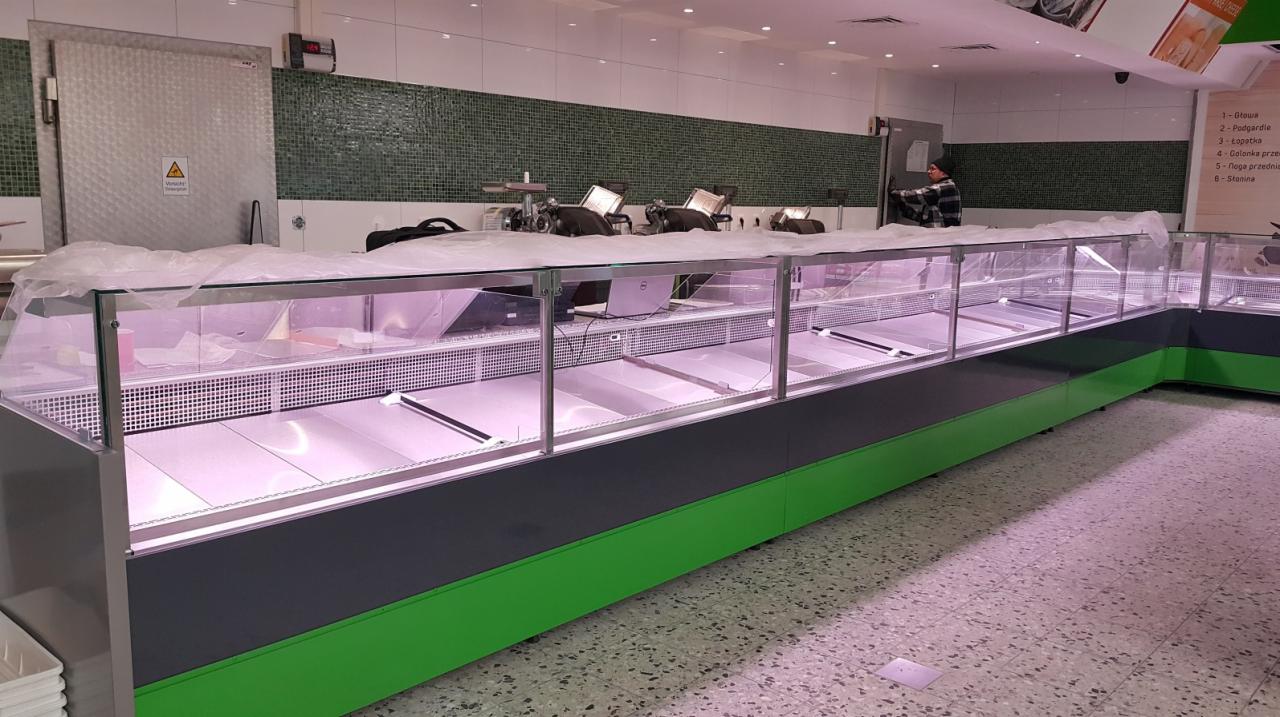 Nowa, pusta lada chłodnicza z zielono-szarym wykończeniem w sklepie, jeszcze zabezpieczona folią ochronną, z widocznymi urządzeniami chłodniczymi na zapleczu.
