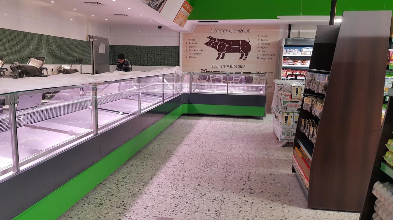 Pusta lada chłodnicza w sklepie mięsnym, z grafiką przedstawiającą podział wieprzowiny i wołowiny na elementy, oświetlenie sufitowe, posadzka lastryko, w tle pracownik sklepu.