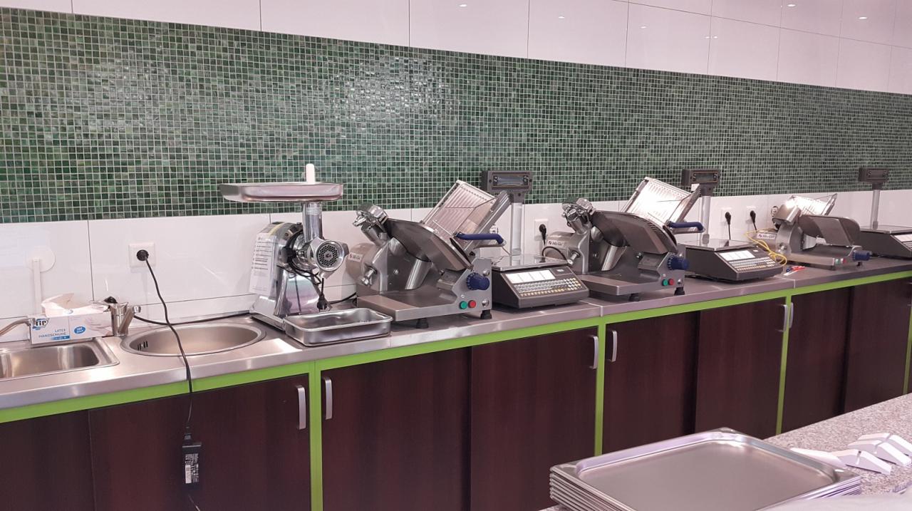 Wyposażenie zaplecza sklepu mięsnego: krajalnice do wędlin, maszynka do mięsa i wagi na blacie z zielonym akcentem, tło z zielonej mozaiki.