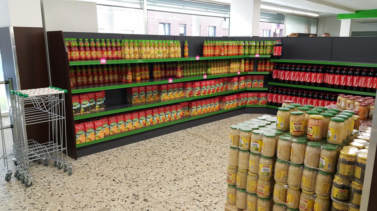 Wnętrze sklepu spożywczego z regałami wypełnionymi napojami i słoikami z przetworami, widoczne wózki sklepowe i podłoga w plamki.