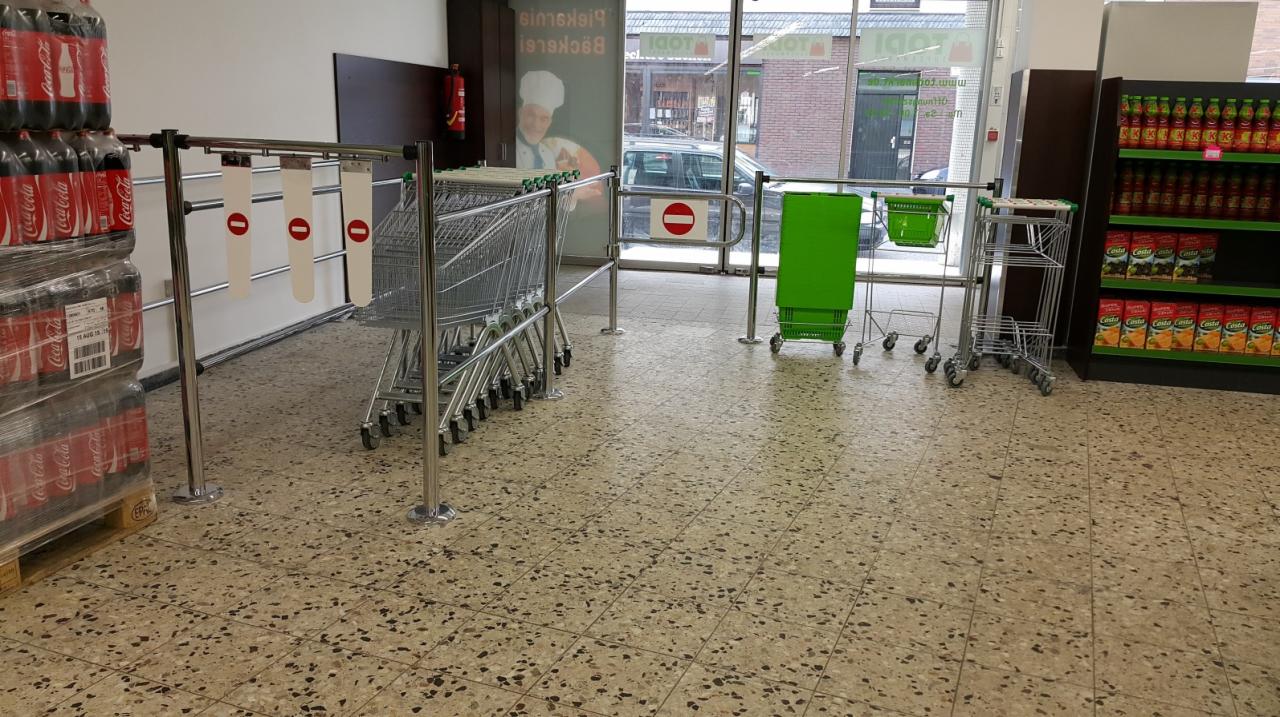 Wnętrze sklepu spożywczego z wózkami, paletą Coca-Coli, regałem z napojami i bramką z zawieszkami z symbolem zakazu wstępu.