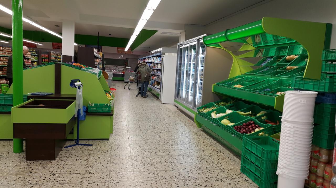 Wnętrze sklepu spożywczego z zielonymi regałami na warzywa i owoce, chłodniami i klientem z wózkiem w tle.