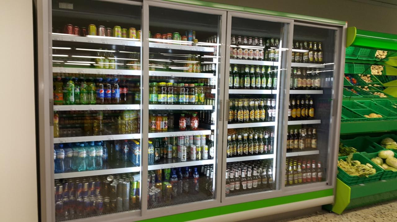 Chłodnia sklepowa z przesuwnymi szklanymi drzwiami, wypełniona napojami w puszkach i butelkach, obok regał z zielonymi pojemnikami na warzywa.
