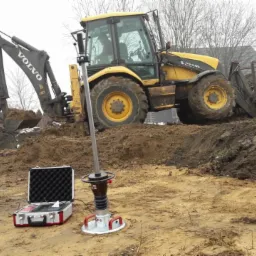 Żółta koparko-ładowarka Volvo BL71 z łyżką wypełnioną ziemią na placu budowy, obok sprzęt do badania zagęszczenia gruntu w walizce transportowej.