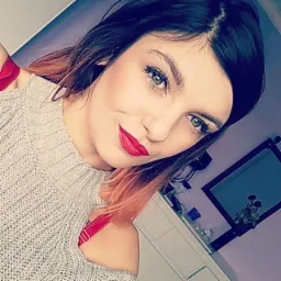 Portret kobiety z czerwonymi ustami, w szarym swetrze z odkrytymi ramionami, patrzącej w obiektyw, z widoczną częścią wnętrza pomieszczenia z fioletową ścianą i komodą.