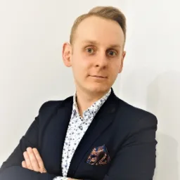 Portret mężczyzny w granatowej marynarce i wzorzystej koszuli, z poszetką w brązowe ornamenty, stojącego na tle białej ściany z założonymi rękoma.