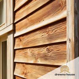 Drewnohouse Piotr Latosiński - Szczegółowe ujęcie elewacji budynku z desek o wyraźnym usłojeniu, z widocznym fragmentem okna i drewnianą ramą. Ciepłe oświetlenie podkreśla naturalną strukturę drewna.