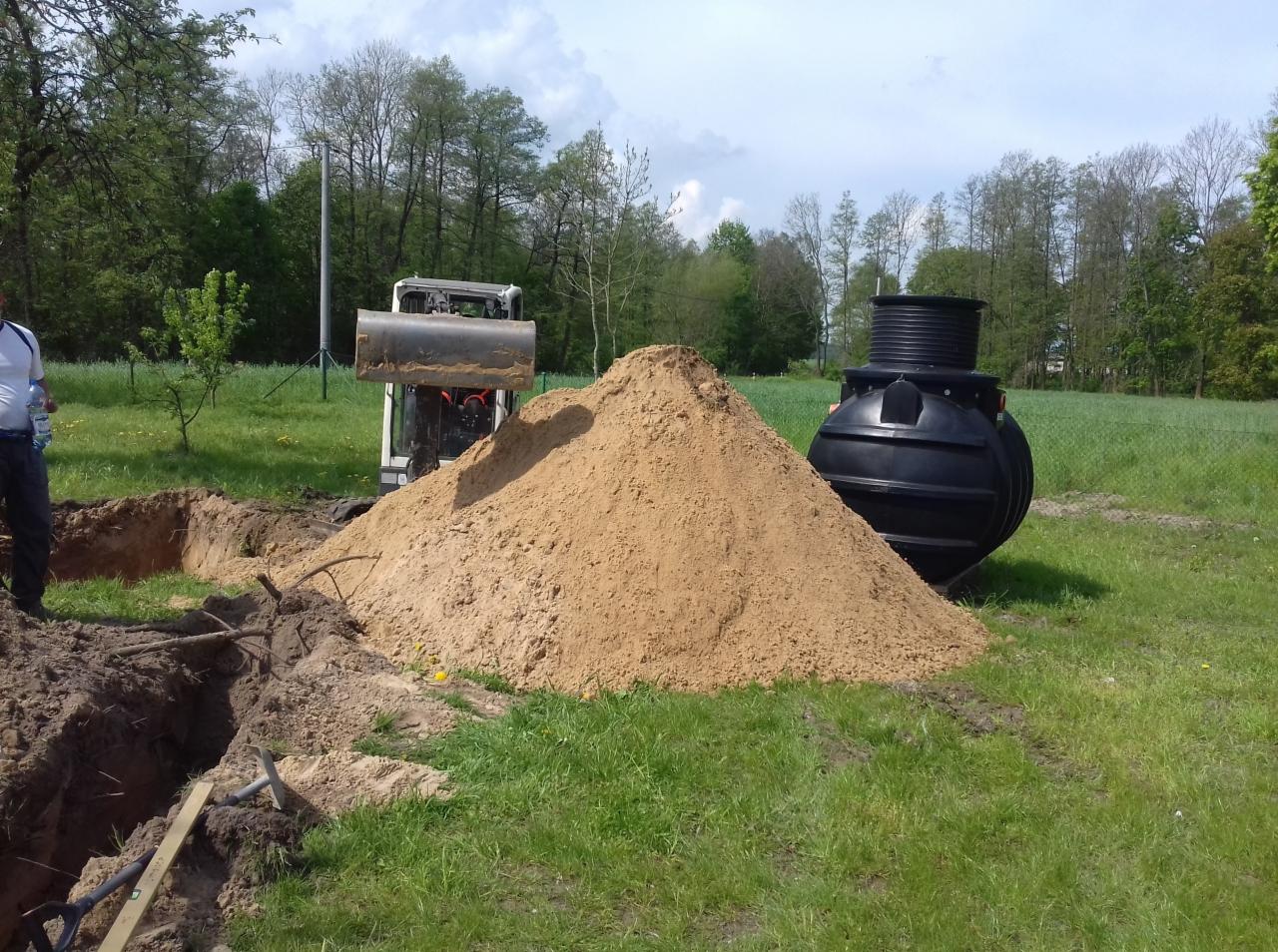 Czarna, plastikowa oczyszczalnia przydomowa obok wykopu i sterty piasku, z mini-koparką w tle na zielonym trawniku.