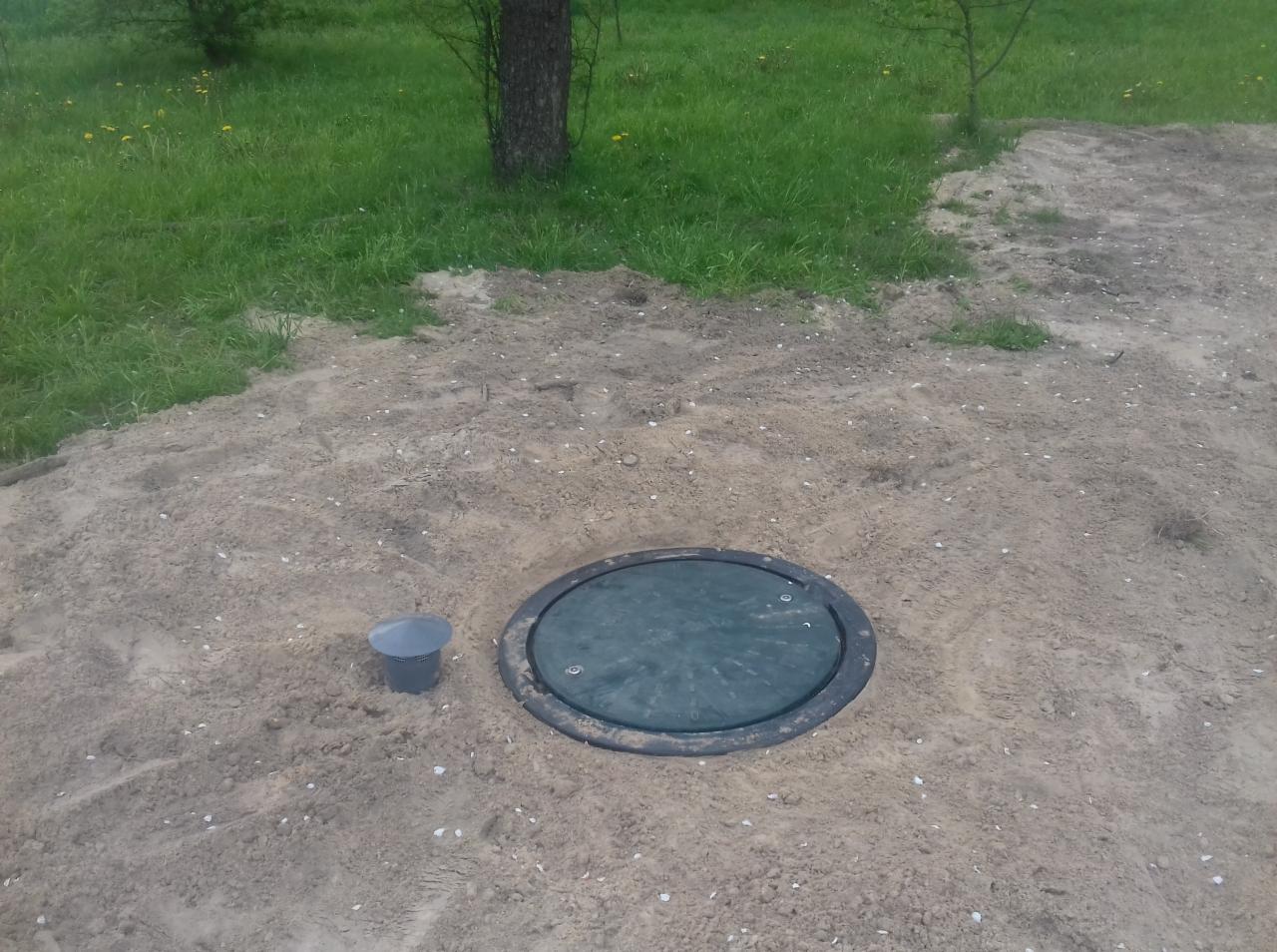 Zielona pokrywa włazu przydomowej oczyszczalni ścieków, częściowo zasypana piaskiem, z widocznym małym, szarym kubełkiem obok, na tle trawy i drzew.