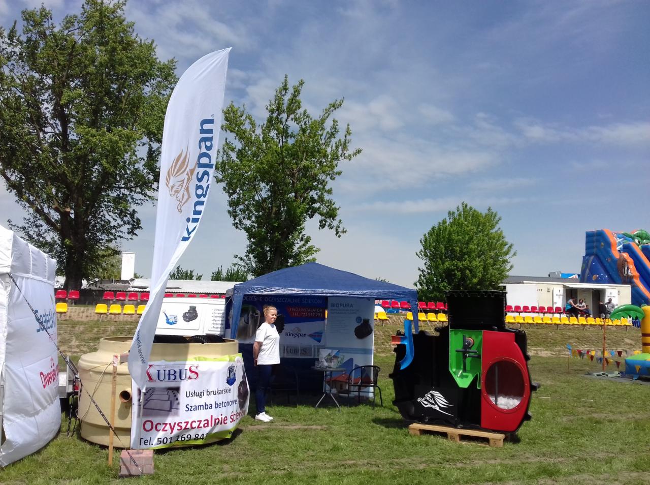 Ekspozycja firmy z Grodziska Wielkopolskiego na targach: stoisko z flagą Kingspan, zbiornikiem na szambo, ulotkami i ekspozytorem oczyszczalni ścieków, w tle dmuchany zamek.