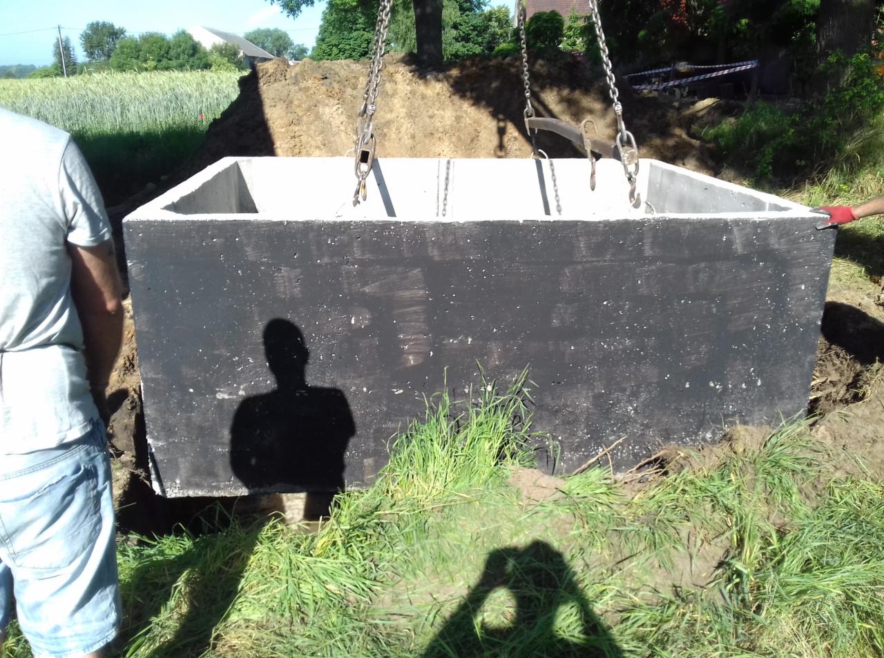 Czarny, betonowy zbiornik opuszczany na łańcuchach do wykopu, widoczne trawy i cień osoby.