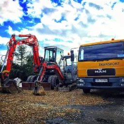 Czerwony minikoparka O&K z dwiema łyżkami i żółta ciężarówka MAN Transport na żwirowym podłożu, w tle las i błękitne niebo z chmurami.