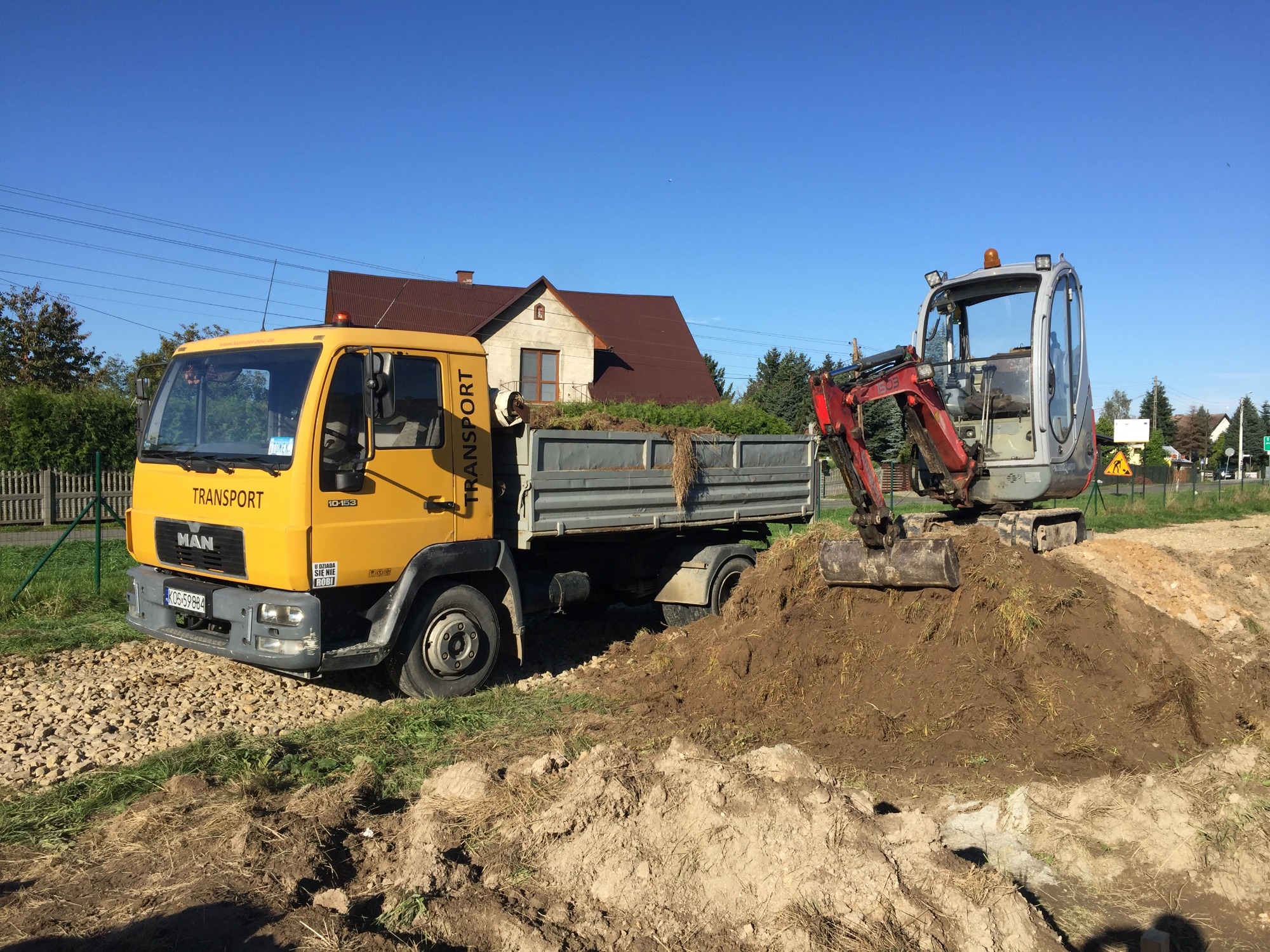 Żółta ciężarówka MAN z wywrotką załadowaną ziemią i mała koparka na hałdzie ziemi na tle domu i błękitnego nieba.