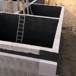 Budowa fundamentów: ściany fundamentowe z bloczków betonowych, izolowane czarną powłoką, widoczna drabina i wykop z hałdami ziemi w tle.