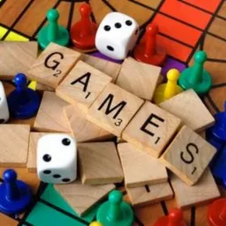 Plansza do gry z pionkami, kostkami i drewnianymi klockami układającymi słowo 'GAMES' w centralnej części kadru.