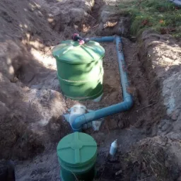 Montaż zielonej, dwukomorowej przydomowej oczyszczalni ścieków z rurami w wykopie ziemnym. Na górnej pokrywie leży wiertarka. Widoczne elementy instalacji.