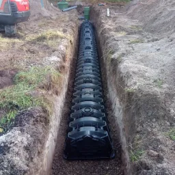 Długa, czarna konstrukcja drenażowa ułożona w wykopie, podsypana kamieniami. W tle widoczny fragment czerwonej koparki Kubota i elementy instalacji.