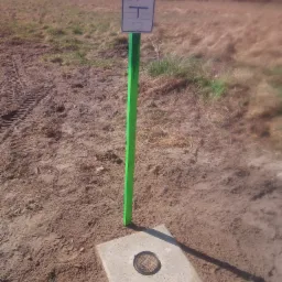 Betonowa studzienka z zielonym słupkiem i tabliczką informacyjną na gruncie, w tle łąka i drzewa. Widoczne ślady opon.