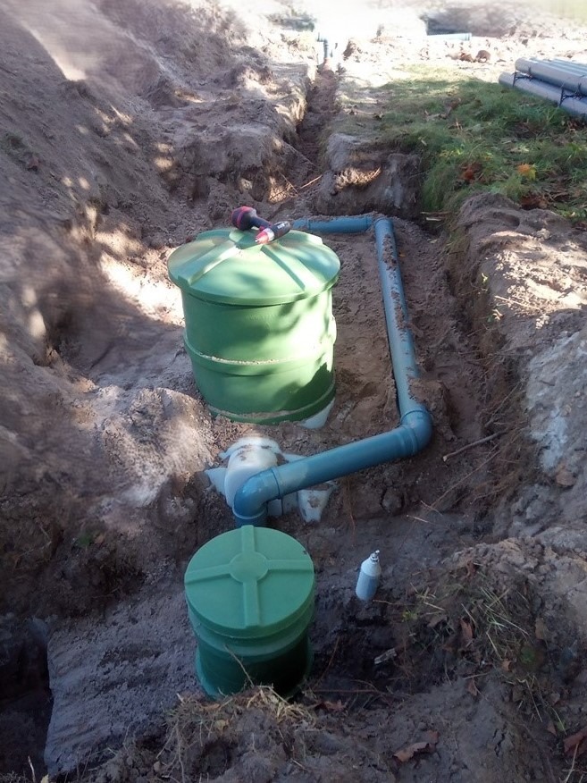 Montaż zielonej, dwukomorowej przydomowej oczyszczalni ścieków z rurami w wykopie ziemnym. Na górnej pokrywie leży wiertarka. Widoczne elementy instalacji.