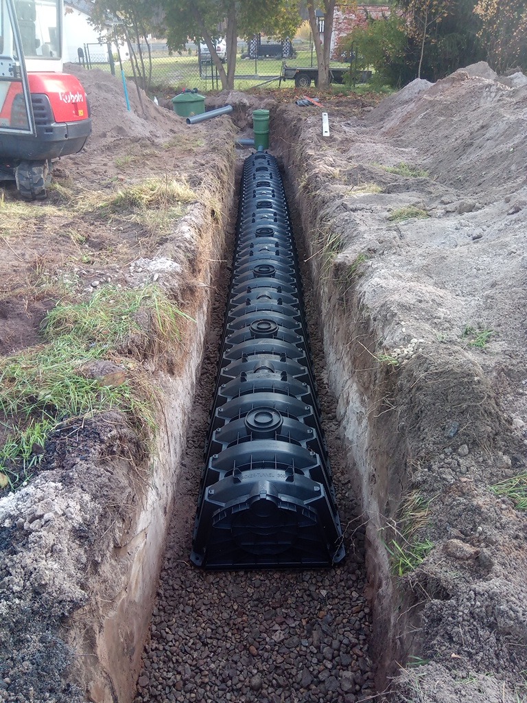 Długa, czarna konstrukcja drenażowa ułożona w wykopie, podsypana kamieniami. W tle widoczny fragment czerwonej koparki Kubota i elementy instalacji.