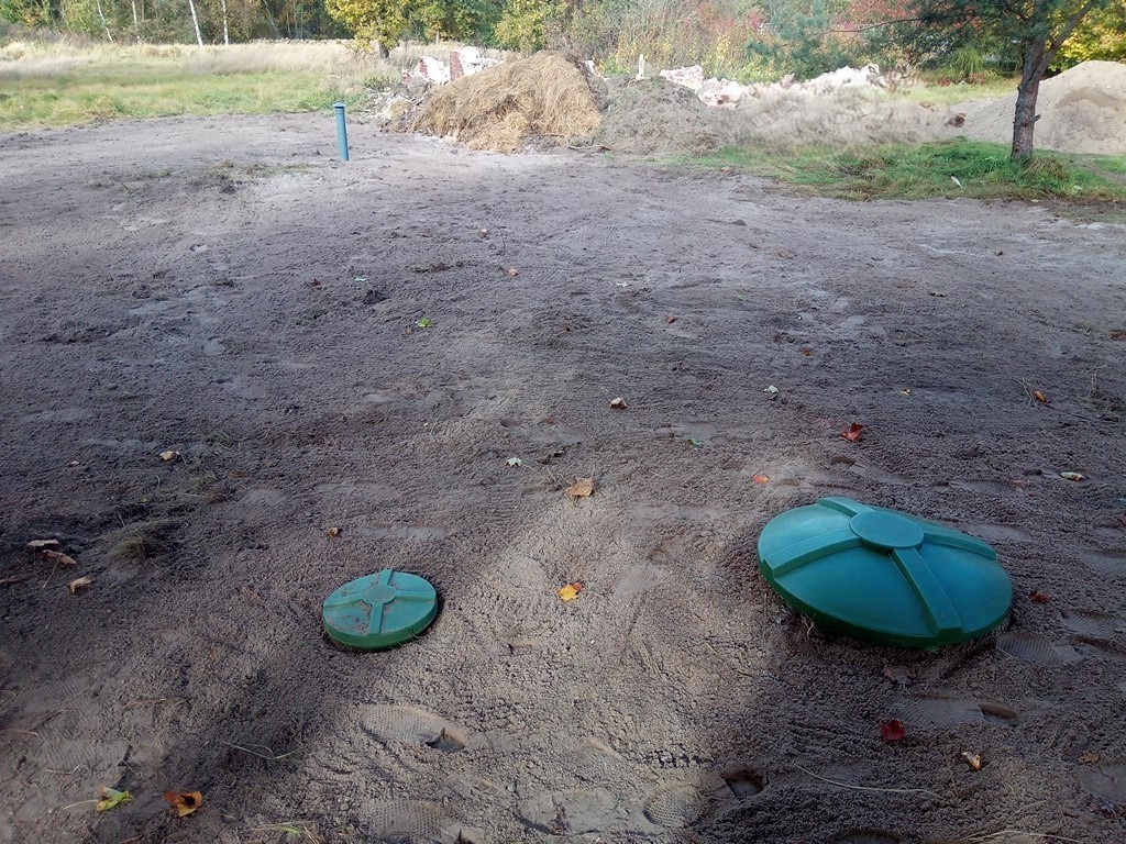 Dwa zielone włazy szamba betonowego na ziemi w Witkowie. W tle widać pryzmę piasku i drzewa. Ślady stóp na ziemi sugerują niedawne prace.