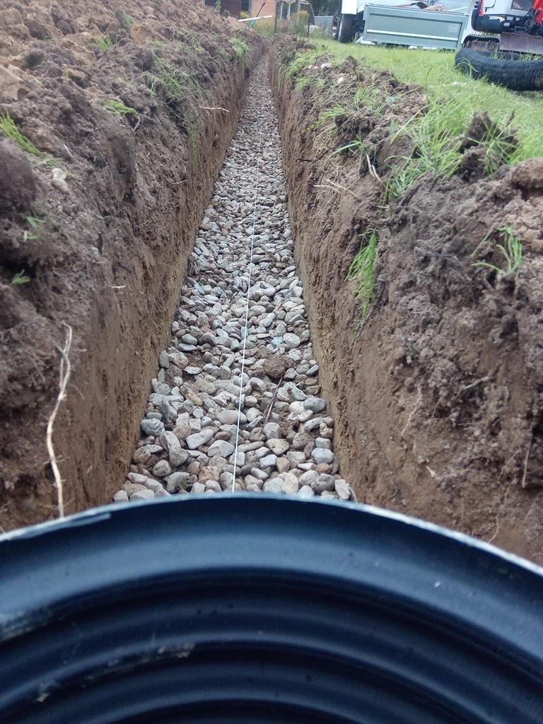 Wykopany rów wypełniony kamieniami, prawdopodobnie drenaż. W tle trawa, ziemia i sprzęt budowlany. W dolnej części widoczny element instalacji.