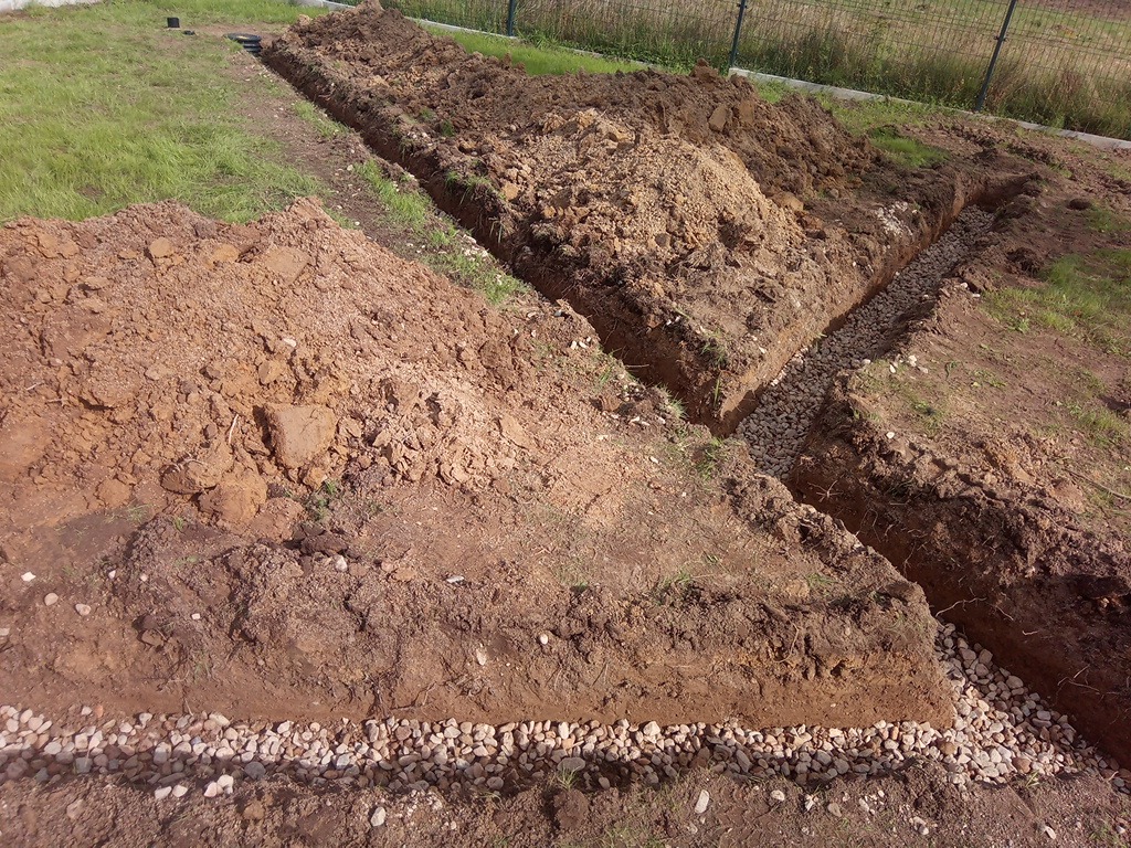 Wykopane rowy w ziemi, częściowo wypełnione kamieniami, prawdopodobnie pod drenaż. Widoczne pryzmy ziemi i trawa. Metalowe ogrodzenie w tle.