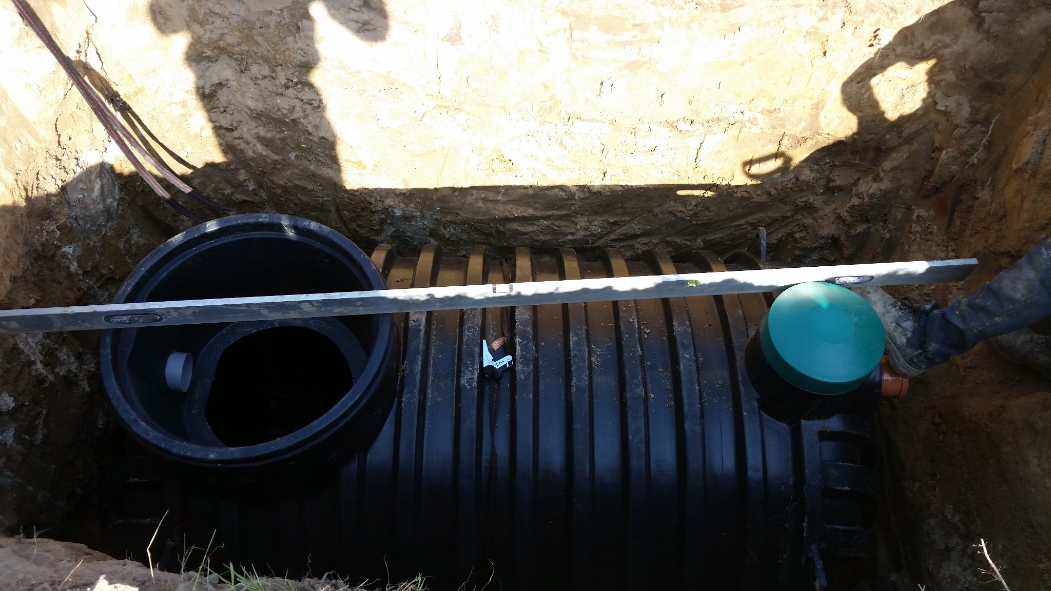 Czarny zbiornik w wykopie, poziomowany. Widoczny fragment wykopu, kable i poziomica. Zielona pokrywa zbiornika. Etap instalacji.