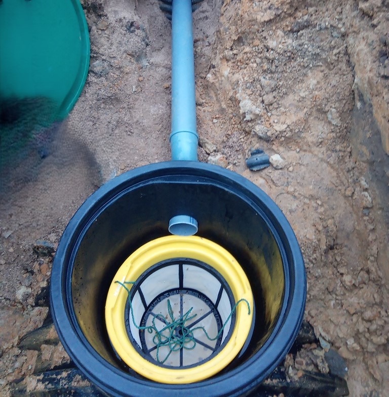 Montaż przydomowej oczyszczalni: czarna studzienka z żółtym filtrem w ziemi, niebieska rura doprowadzająca, widoczny fragment zielonej pokrywy.