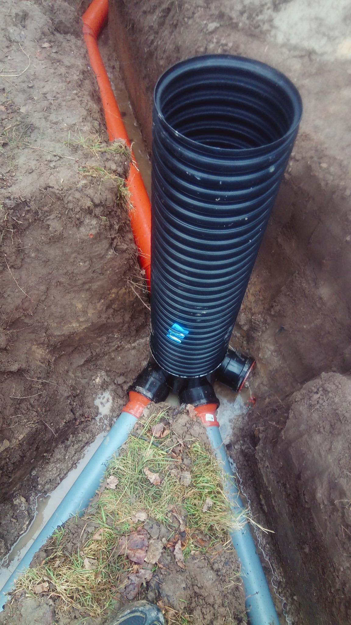 Studnia drenażowa z podłączonymi rurami w wykopie. Widoczne połączenia i fragmenty rur w ziemi.