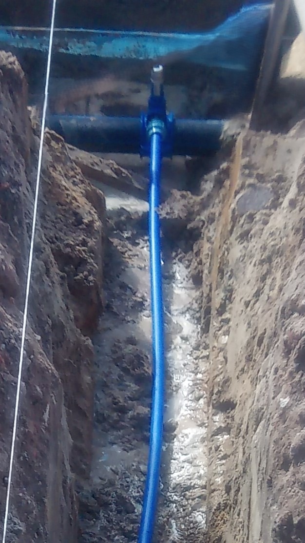Niebieska rura pionowa w wykopie ziemnym z zaworem, podłączona do rury poziomej. Widoczne ślady wilgoci i błota, sugerujące świeżo wykonane prace instalacyjne.