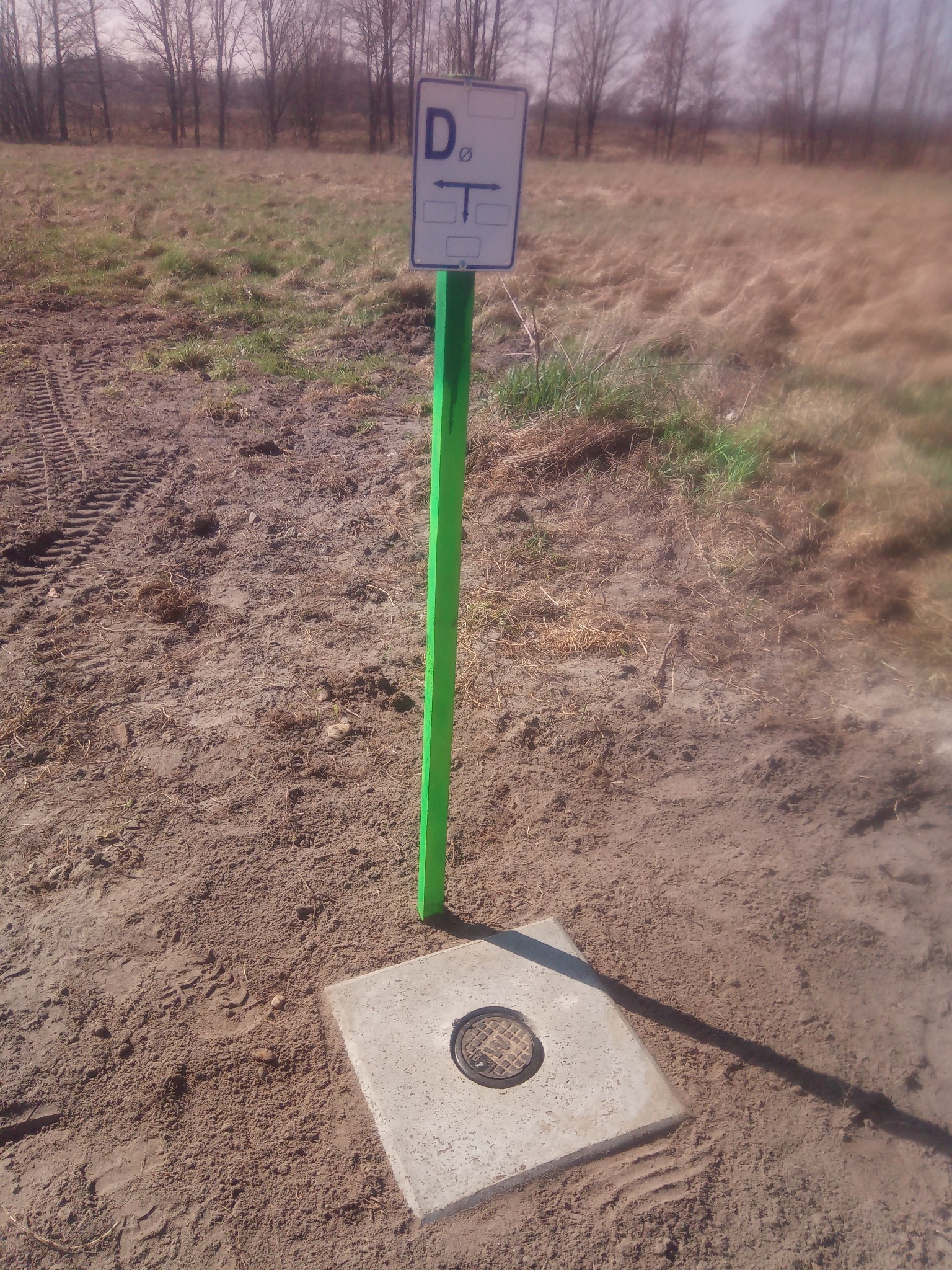 Betonowa studzienka z zielonym słupkiem i tabliczką informacyjną na gruncie, w tle łąka i drzewa. Widoczne ślady opon.