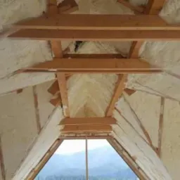 Poddasze domu po ociepleniu pianką poliuretanową, widoczne drewniane elementy konstrukcyjne i okno z widokiem na las.