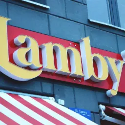 Zewnętrzny szyld z nazwą 'Lamby's' w kolorze żółto-białym na czerwonym tle, zamontowany na szarej ścianie budynku, z widocznym fragmentem markizy w biało-czerwone pasy i tabliczką z nazwą ulicy 'Ul...
